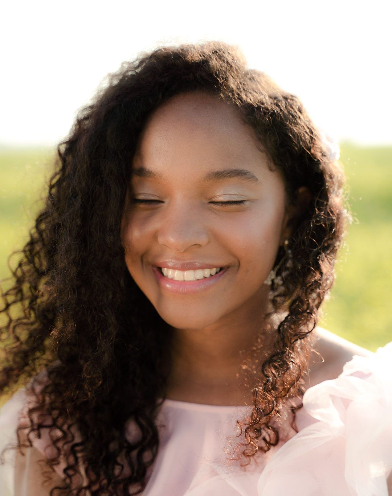Une jeune femme aux cheveux bouclés sourit au soleil, les yeux fermés.